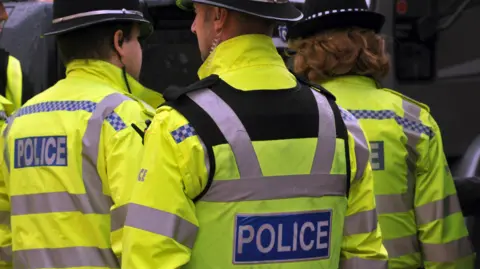 A rear view of three West Midlands Police officers - two male and one female - all wearing bright yellow high-vis police jackets and wearing helmets. 