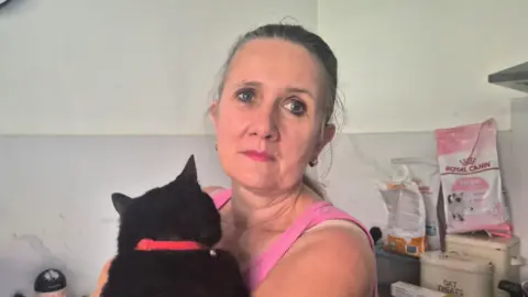 BBC A woman holding a black cat whilst wearing a pink vest, lookintg at the camera.