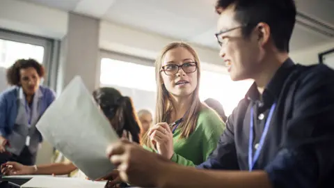 Getty Images Two students look at each other and converse while holding up pens and paper in a university seminar, with a lecturer and other students in the background slightly out of focus