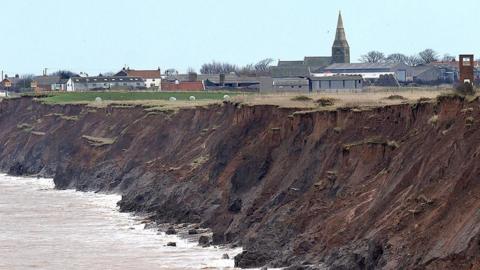 Brown cliff edge with the sea lapping up against it and settlement in the far distance