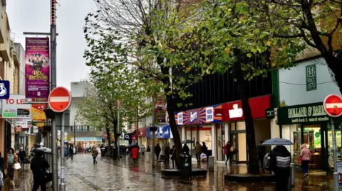 A street on a rainy day. Pedestrians are holding umbrellas as they walk past high street shops.