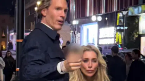 British Transport Police A CCTV image showing a tall man with brown hair wearing wearing a smart dark blue jacket, gesturing forward. He is with a woman with long, blonde hair 