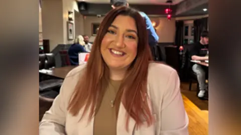 A woman looking at the camera. She has long brown hair and smiles at the camera. She is wearing a beige blazer with a brown top underneath.