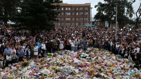 Reuters A huge crowd of people stands at a vigil next to the beach in Bondi. There are several large trees in the background and a large brick building. In the foreground are hundreds of floral tributes carpeting the ground. 