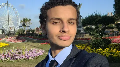 Contributed A selfie style picture of Alkhatip who has short curly black hair and is wearing a dark suit with a blue shirt. He is standing in the cliff top gardens in Clacton with a ferris wheel in the background.