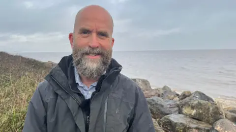 Fayers stood in front of rocks near to the sea. He is wearing a grey jacket and has a grey beard.