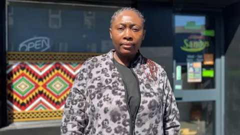 Shows a woman with her hair in plaits and a cardigan standing in front of a restaurant 