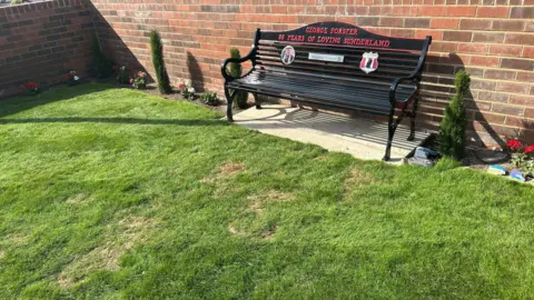 Black metal memorial bench positioned against a red-brick wall in front of a well-manicured patch of grass. Red lettering on the bench reads, “GEORGE FORSTER” and “99 YEARS OF LOVING SUNDERLAND". You can also see the red and white logo of Sunderland AFC. There are flowers planted in the ground on the edge of the walling.