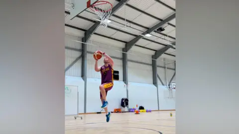 Tiverton Titans A player leaps in the air with both feet off the ground. A basketball hoop is above him and he is wearing a purple and yellow kit.