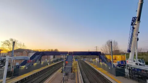 Network Rail A crane on a station platform next to a newly installed blue footbridge which is connecting the platforms. The sun is rising in the background.