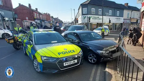 A police car which says 'interceptor' on it is parked diagonally across a road next to a black car which appears to have driven into black railings on a pavement. A police motorbike and police officers are behind the cars, which are on a busy road with shops.