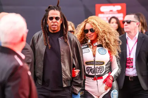 Getty Images Picture of Jay-Z wearing a brown jacket with a t-shirt underneath and Beyoncé wears a white racing suit with red and black accents.