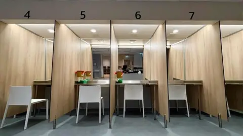 Numbered booths with a white chair in each booth. Each one faces a mirrored wall. It is brightly lit.