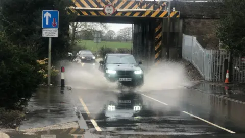 A car can be seen driving through flood water under a railway bridge. Water can be seen spraying on either side of the vehicle as it drives through. There are cars waiting to go through behind it. 