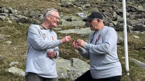 Steve Egan Ivor Ramsden (left) wearing a grey sweatshirt and glasses, passing the dog tag to Don Madar who is also wearing a grey sweatshirt with a black baseball cap. They're on the rocky hillside and there is a white flag pole to the right.