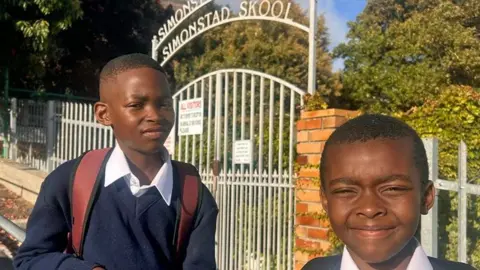Brothers Anele (left), who is carrying a rucksack, and Lifalethu Mbasana in their navy jumper and white shirt uniforms outside their school gate in Simon's Town. Incorporated into the the white-painted metal gate is a curved sign reading 'Simonstad Skool'