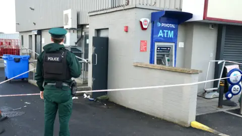 A police officer, in uniform, is standing watch outside of the damaged ATM. A door to the left of the machine is open and police tape surrounds it. The ATM is bright blue in colour. 