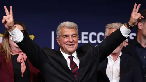 Joan Laporta raises his hands in celebration after winning the Barcelona presidency election. He has grey hair and is wearing a dark suit, white shirt and a tie in Barcelona's club colours - red and blue.