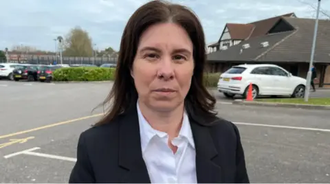 Leanne Reynolds looking at the camera standing in a car park. She has dark shoulder-length hair and is wearing a white shirt and a dark-coloured jacket