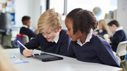 Getty Images A blonde boy and a girl with black hair sit at a desk with a tablet, the boy is using a white pen to operate the tablet. They both look at the screen with interest. They are both wearing navy school uniform