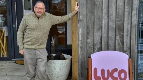 LDRS Bernie Smith is wearing all grey. He is standing outside of a wood clad building which is a pilates studio. In the foreground is a pink sign saying LUCO which is the studios brand name. 