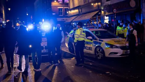Getty Images Several police officers standing near two police cars with blue flashing lights outside a strip of bars. There are lots of people standing around in the road.