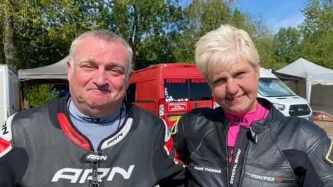 BBC Dave and Alison looking into the camera and smiling. They're both wearing black motorbike leathers. Dave has short grey hair with sideburns and is wearing a blue undershirt. Alison has short blonde hair and is wearing a pink undershirt. They're stood in front of their red van, which has 'Terrier Racing' printed on the rear window. 