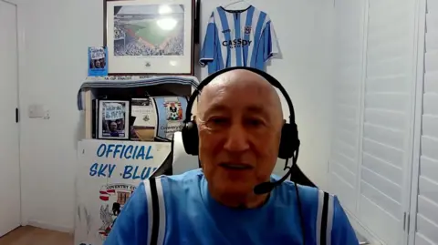 John Mullaney sits with Coventry City paraphernalia behind him and earphones and a speaker set. He is wearing a Coventry City top
