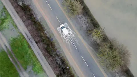 PA Media An aerial view shows a white car driving down a road with floodwater in the road and beside it, in Leicestershire on Monday.