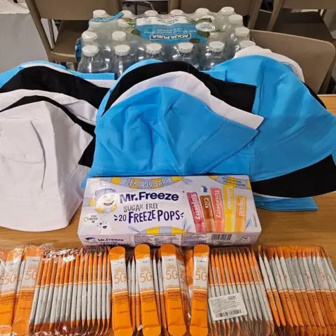 Good Shepherd A pack of water bottles, several caps, ice lollies and orange hydration sachets on top of a table.