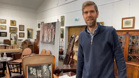 Edd Smith/BBC A man standing dressed in blue stood adjacent to the suitcase of memorabilia 