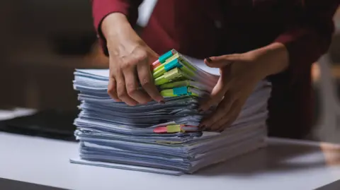 Getty Images A pile of documents is stacked on a desk and a woman's hands are seen sorting through them.