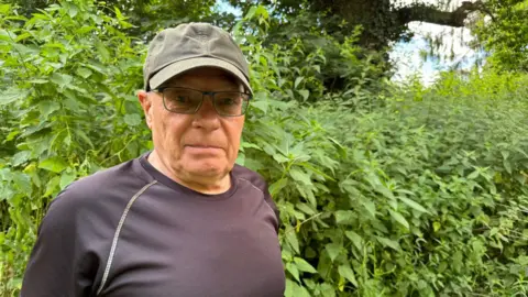 A middle-aged man wearing a black t-shirt, glasses and green baseball cap. He is stood in front of a large bush of stinging nettles and is looking into the camera.