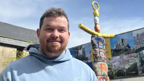 The image shows a man with brown hair and a ginger beard wearing a light blue hooded sweatshirt looking directly at the camera. He is standing outside with the brightly-coloured statue behind him. The sky is blue and in the background are tall buildings.