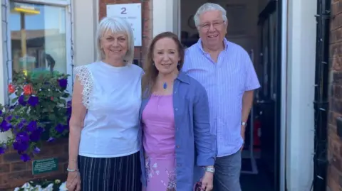 BBC/Angela Ferguson A woman with shoulder length grey hair and a short-sleeved white blouse and blue and white striped trousers is standing next to a woman with long brown hair, a pink top and skirt and a denim shirt and a man wearing a light blue short sleeved pin stripe shirt and jeans. All are smiling while standing in front of the entrance to the Station Master's House. There are flowers to the left.