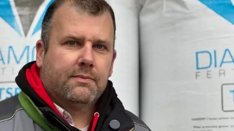 Andrew Sinclair/BBC Russel Abbott standing in front of bags of fertiliser. He has a beard and is wearing a grey coat.