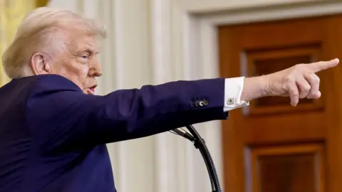 Getty Images Profile view of Donald Trump, wearing a navy suit, pointing his figure during a press conference