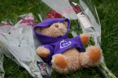 A teddy bear in a purple hoodie lies on grass, next to bunches of wrapped flowers