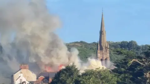 Colin McGrath A horizon of the tops of buildings and green trees. A towering grey church spire sits above the rest of the buildings. Grey smoke is bellowing from the trees to the left of the church. 