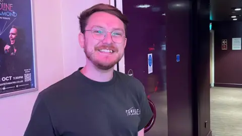 Matt Knight/BBC A man wearing a black top and glasses while smiling at the camera. There is a poster behind him and he is standing by a door.
