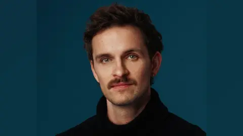 Rebecca Need-Menear Vittorio looking at the camera. He is wearing a black jumper. He has brown hair and green eyes. The background is a blue/green colour.