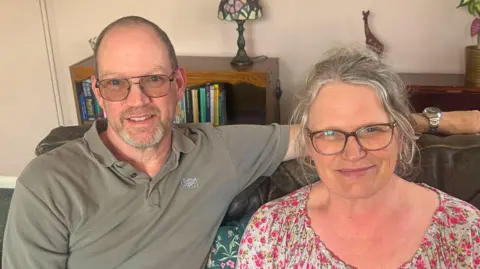BBC/ Emily Johnson A man with glasses and a grey t-shirt puts his arm around a woman in a floral top. They sit on a sofa in a living room.