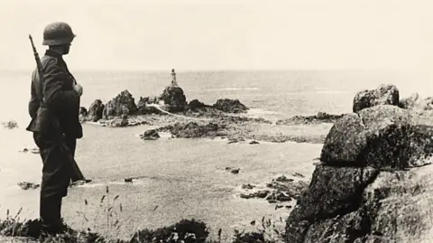 Getty Images A black and white photo of a German soldier holding a rifle and wearing uniform and a tin helmet is standing on a cliff and looking out to sea at Corbiere. There is a rocky outcrop with a lighthouse.