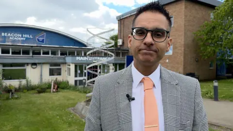 A man wearing glasses, patterned blazer, and peach tie with lapel microphone standing outside Beacon Hill Academy main entrance, with a spiral sculpture and school buildings in the background.
