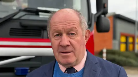 George King/BBC A head and shoulders image of Tim Passmore. He is wearing a navy blazer over a blue jumper, which is over a pink shirt with a tie. He is looking directly into the camera. 