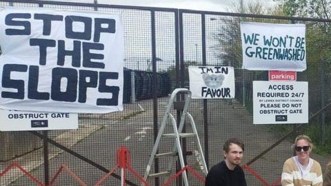 A man and woman crouch down in front of metal gates emblazoned with banners reading "stop the slops", "we won't be greenwashed" and pictures of rats