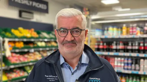 A person wearing glasses and a dark jacket stands facing the camera inside a small grocery store. Shelves filled with fresh produce, including bananas and other fruits and vegetables, are visible on the left, while refrigerated drink shelves with bottles and cans line the right side. Overhead, promotional signs hang from the ceiling, and a sign reading “Freshly Picked” is visible in the background.
