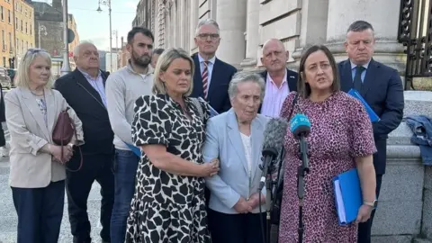 A group of nine people are gathered on a street outside a building - there are six standing behind three women. Of those three, two younger women flank an elderly woman. The three are standing in front of two news microphones. It is during he daytime and a city streetscape can be seen in the background. 