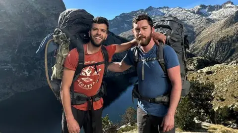 Aziz Ziriat, left, and Samuel Harris in shorts, t-shirt wirth large backpacks in front of a lake surrounded by mountains