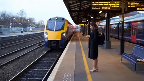 A woman is on her phone while she waits on a train platform. There is a yellow and blue train on the tracks in front of her